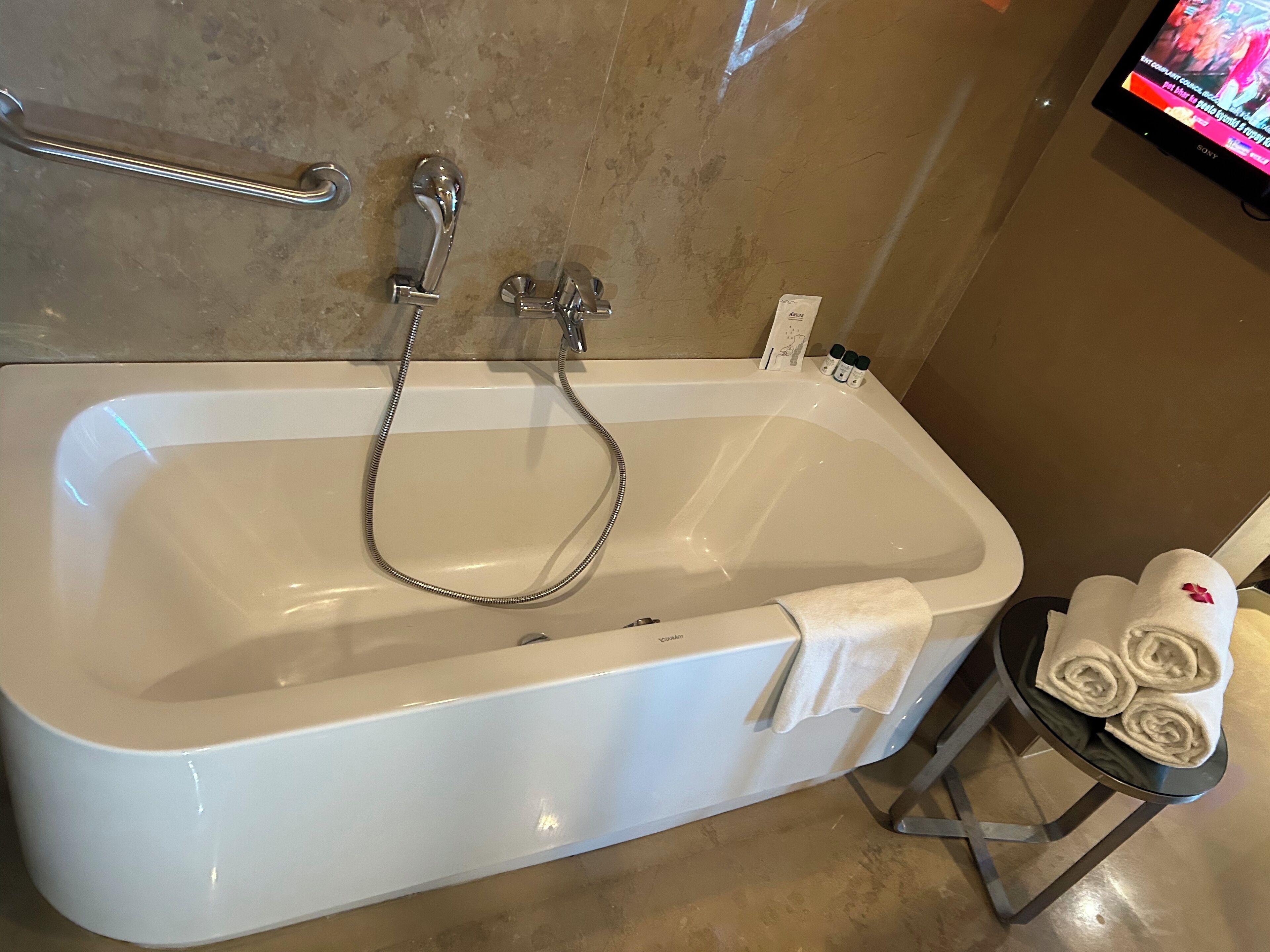 A bathroom with a white bathtub, a handheld showerhead, a towel rack with towels, and a small table with rolled towels.