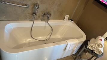 A bathroom with a white bathtub, a handheld showerhead, a towel rack with towels, and a small table with rolled towels.
