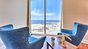 Ein Zimmer mit zwei blauen Stühlen, einem Holztisch und Blick auf den Strand durch ein großes Fenster.