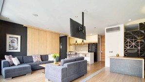 A modern living room with grey sofas, a wooden accent wall, and a kitchen area with a black refrigerator and white cabinets. - ONE BEDROOM HUGE BALCONY IN ENERGY ROOF POOL (Medellin)