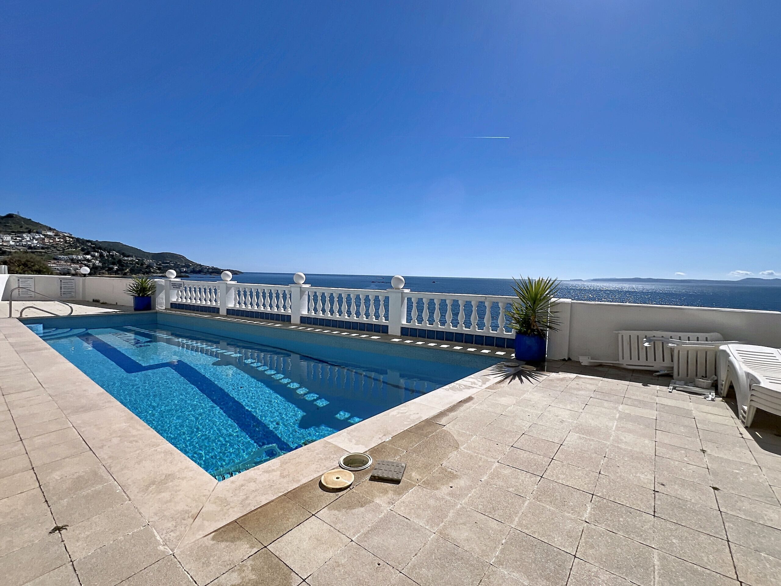 A swimming pool with a view of the sea, a white railing, and a tiled deck.