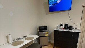 Eine kompakte Küchenzeile mit Mikrowelle, Kaffeemaschine und kleinem Kühlschrank. Eine Bank steht vor einem weißen Tisch mit Spüle. Ein Fernseher ist über der Küchenzeile an der Wand montiert.