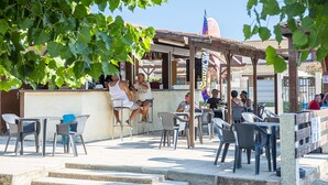 An outdoor café with tables and chairs, a pergola, and a view of a building with a flag. - Portiragnes Group Stay with 2 pools (Portiragnes)