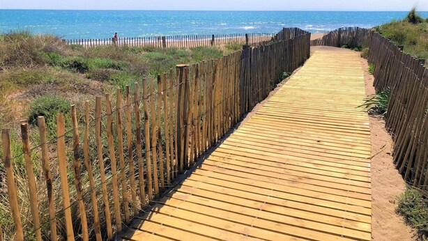 Une promenade en bois mène à une plage, avec une clôture d’un côté et une zone herbeuse de l’autre.