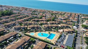 Un quartier résidentiel en bord de mer, doté d’une piscine centrale, entouré de logements aux façades uniformes et offrant une vue dégagée sur la mer.
