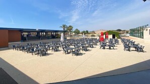 Un espace extérieur aménagé avec de nombreuses tables et chaises, une scène couverte et un bâtiment doté d’une façade en verre.