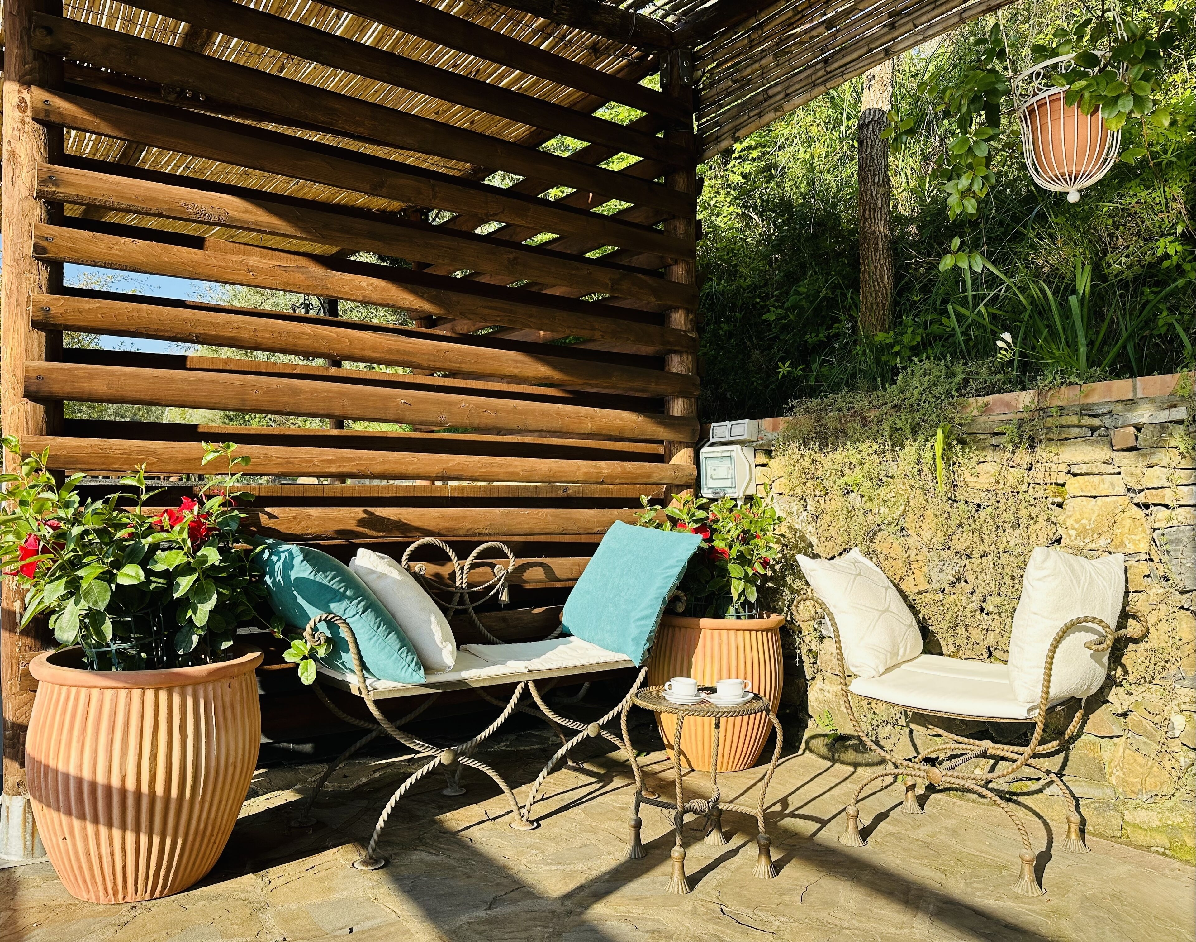 A wooden pergola with a seating area featuring two chairs and a table, surrounded by potted plants and a stone wall.