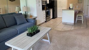 En kompakt opholdsstue med en sofa, et lille bord med en plante, et tekøkken med køleskab, ovn og mikroovn, samt et afsat spiseområde med barstole.