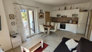 Et moderne køkken med hvide skabe, en mikrobølgeovn og komfur. Der er et spiseområde med et bord og stole. En sort sofa og et fjernsyn er synlige i opholdsstuen.
