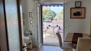 Et værelse med en skydedør, der fører ud til en udendørs terrasse med et bord og stole. En trædør er på højre side, og et lille bord med en stol er på venstre side.