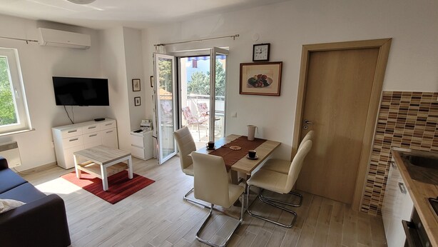 En moderne stue med sofa, fjernsyn, spiseplads med stole og bord samt et køkken med vask og komfur.