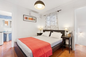 A bedroom with a bed, bedside tables, a nightstand, a wall-mounted air conditioner, a framed picture, and a bathroom visible through an open door.