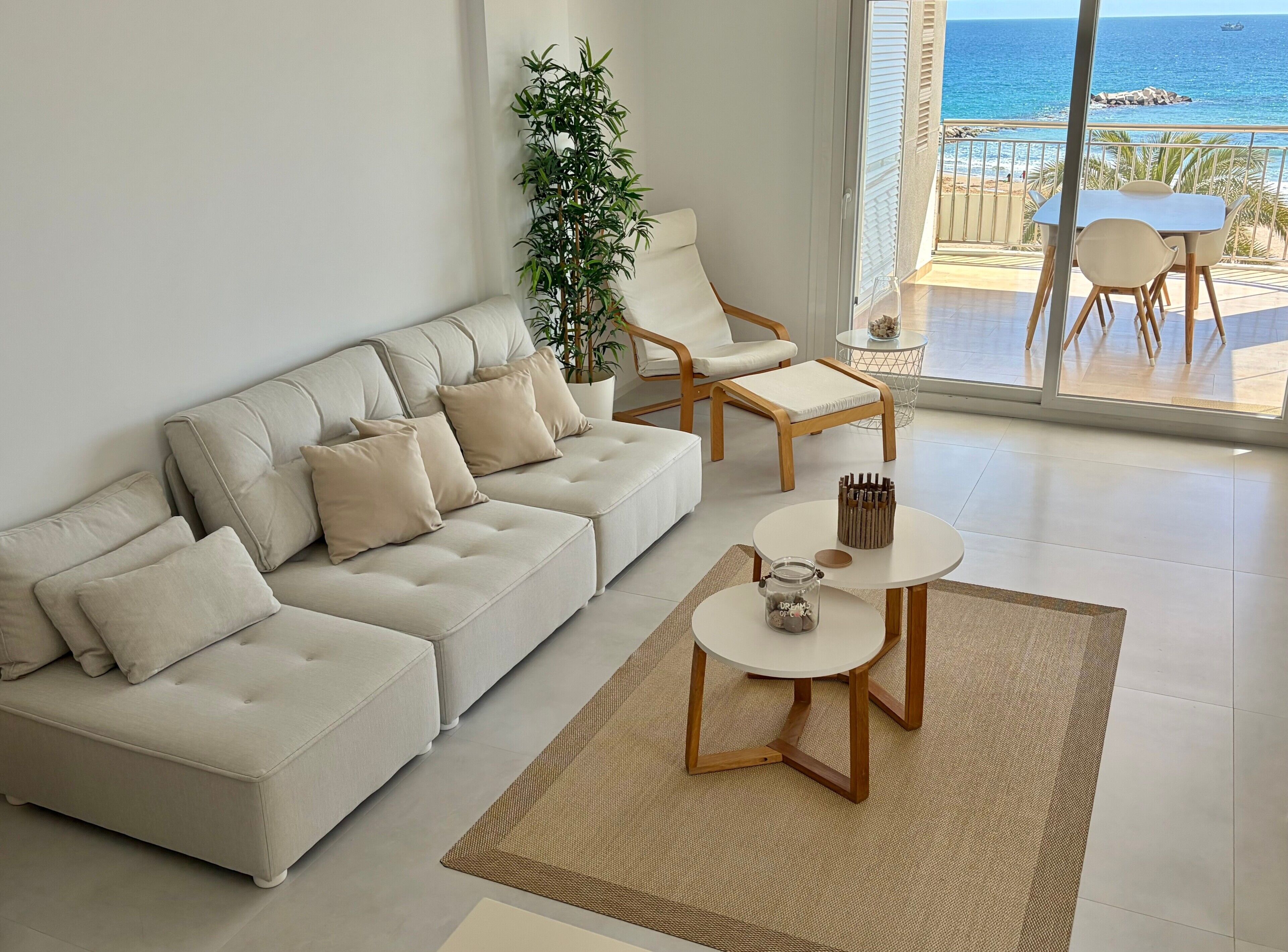 A modern living room with a sofa, armchair, and coffee table, a potted plant, and a view of the sea through a glass door.