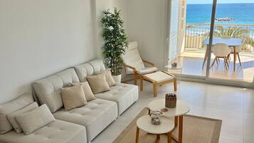 A modern living room with a sofa, armchair, and coffee table, a potted plant, and a view of the sea through a glass door.