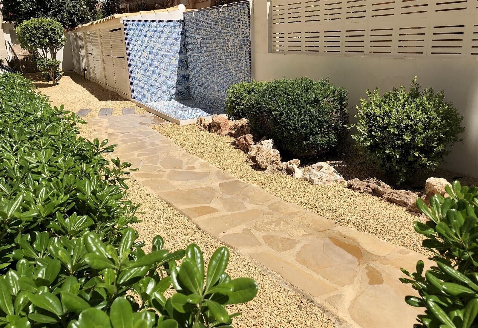 Un sendero de piedra conduce a una zona de piscina con baldosas, rodeada de vegetación y un edificio de fachada color beige.