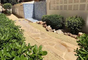 Un chemin de pierre mène à une zone de piscine carrelée, entourée de verdure, et à un bâtiment à la façade beige.