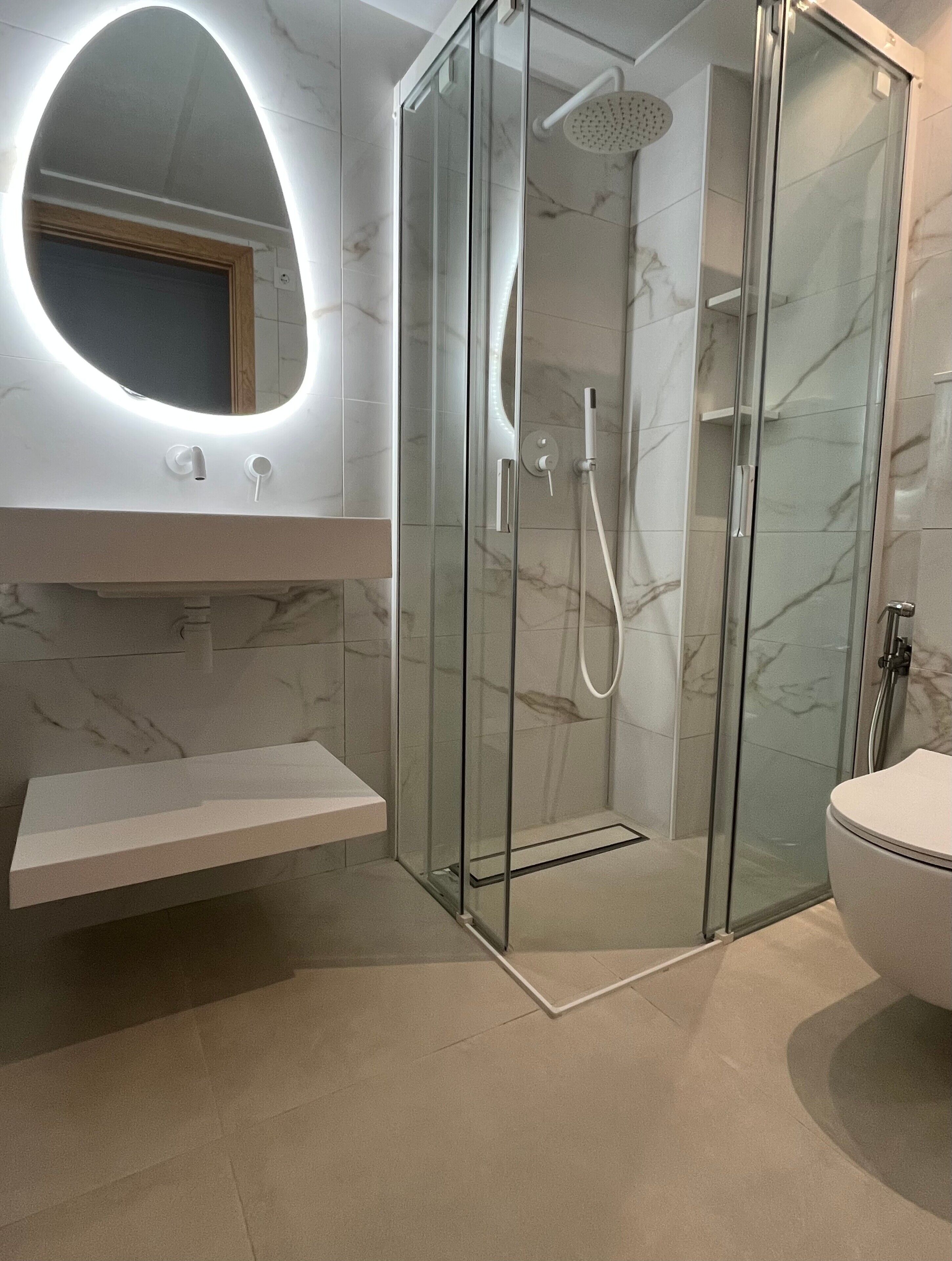 Une salle de bain moderne dotée d’une douche à parois de verre, d’un meuble-lavabo recouvert de carreaux de marbre et d’un grand miroir rond avec éclairage intégré.