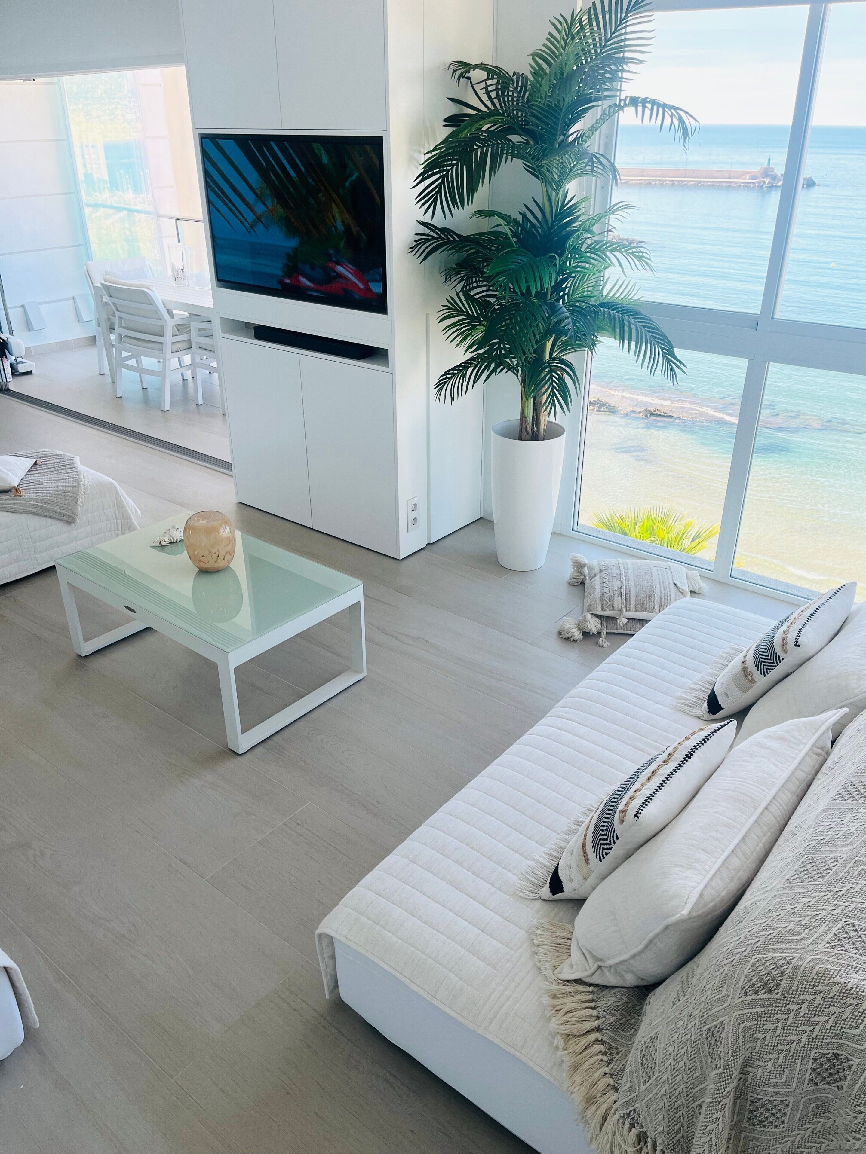 Una sala de estar moderna con un televisor grande, una mesa de centro de vidrio, un sofá blanco, una planta en maceta y vista al mar.