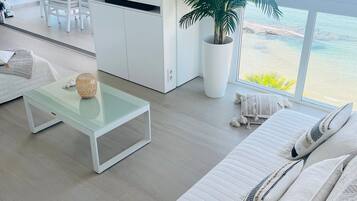 Una sala de estar moderna con un televisor grande, una mesa de centro de vidrio, un sofá blanco, una planta en maceta y vista al mar.