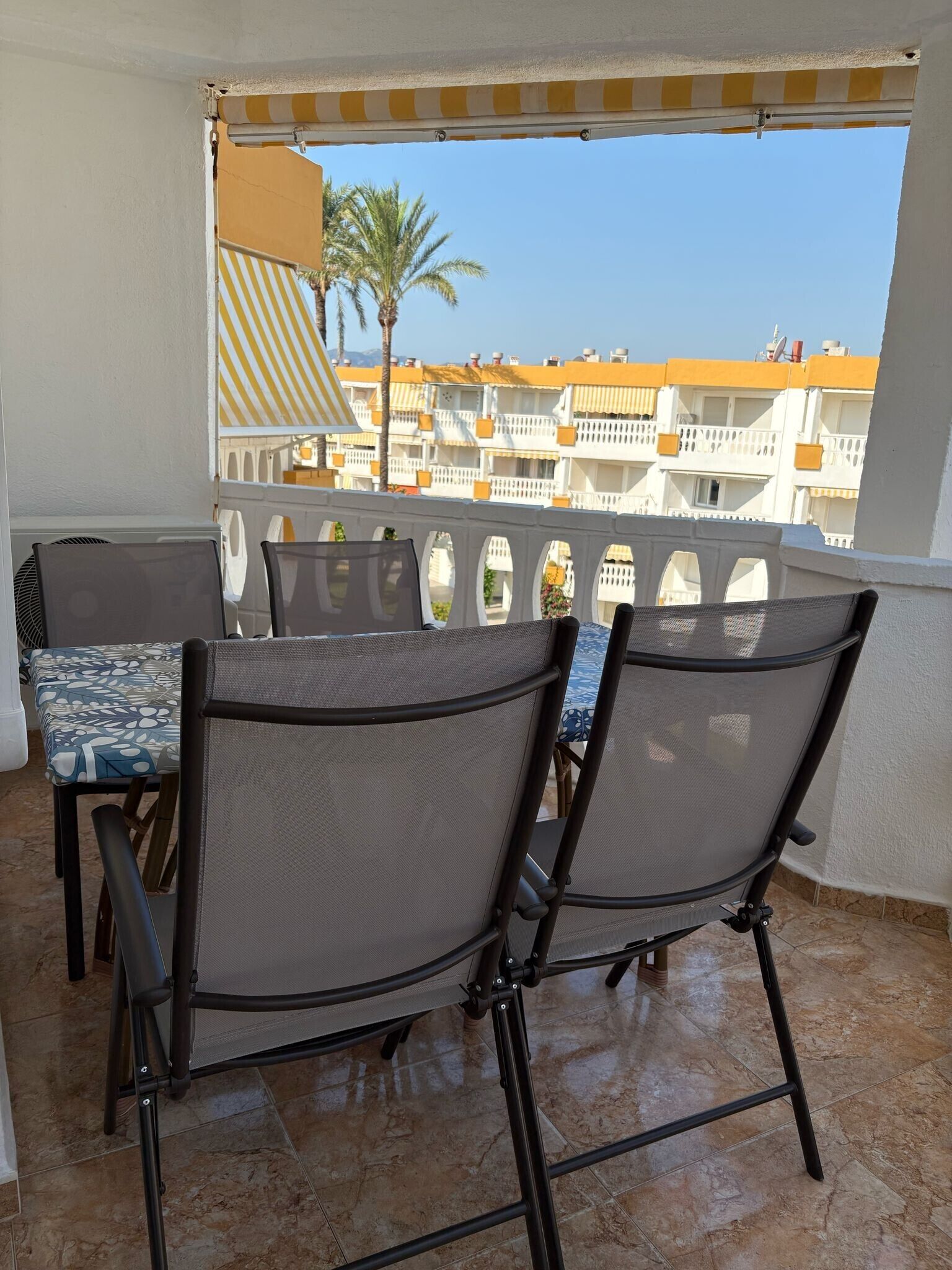 Un balcón con dos sillas y una mesa, un toldo a rayas y vistas a edificios con balcones.