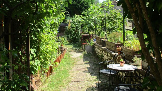 A garden pathway leading to a seating area with a table and chairs, surrounded by lush greenery and a stone wall. - Appartement en sous-sol.Jardin privé. Situé dans un village des montagnes noires (Labastide-Rouairoux)