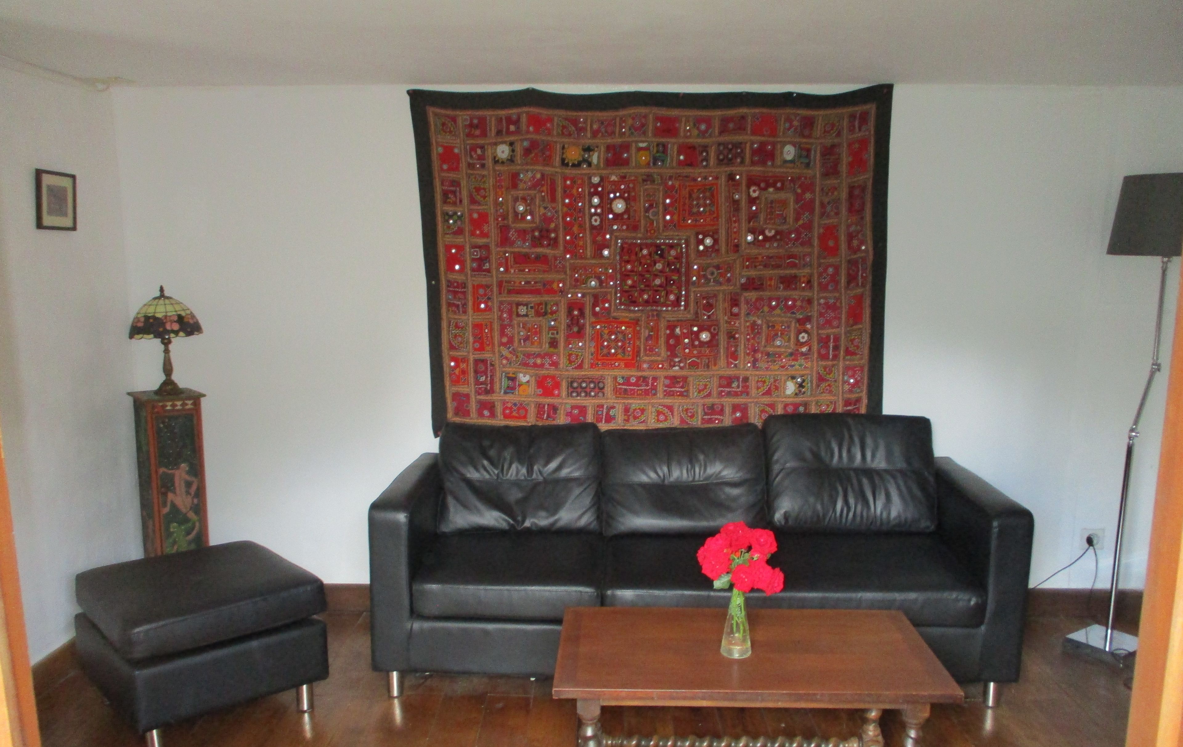 A living room with a black leather sofa, a wooden coffee table with a vase of red flowers, a patterned tapestry on the wall, and a tall floor lamp.