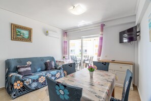 Ein Wohnzimmer mit Sofa, Esstisch, Stühlen, Fernseher und einem Balkon mit Vorhängen.