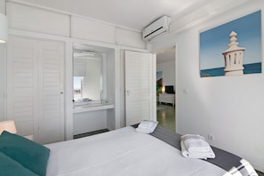 Une chambre d’hôtel avec un lit, un miroir dans la salle de bain, un tableau représentant un phare et une porte donnant sur une autre pièce.
