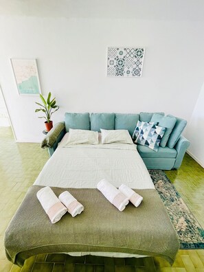 A neatly arranged bedroom with a bed, a sofa, a potted plant, and a framed artwork on the wall.