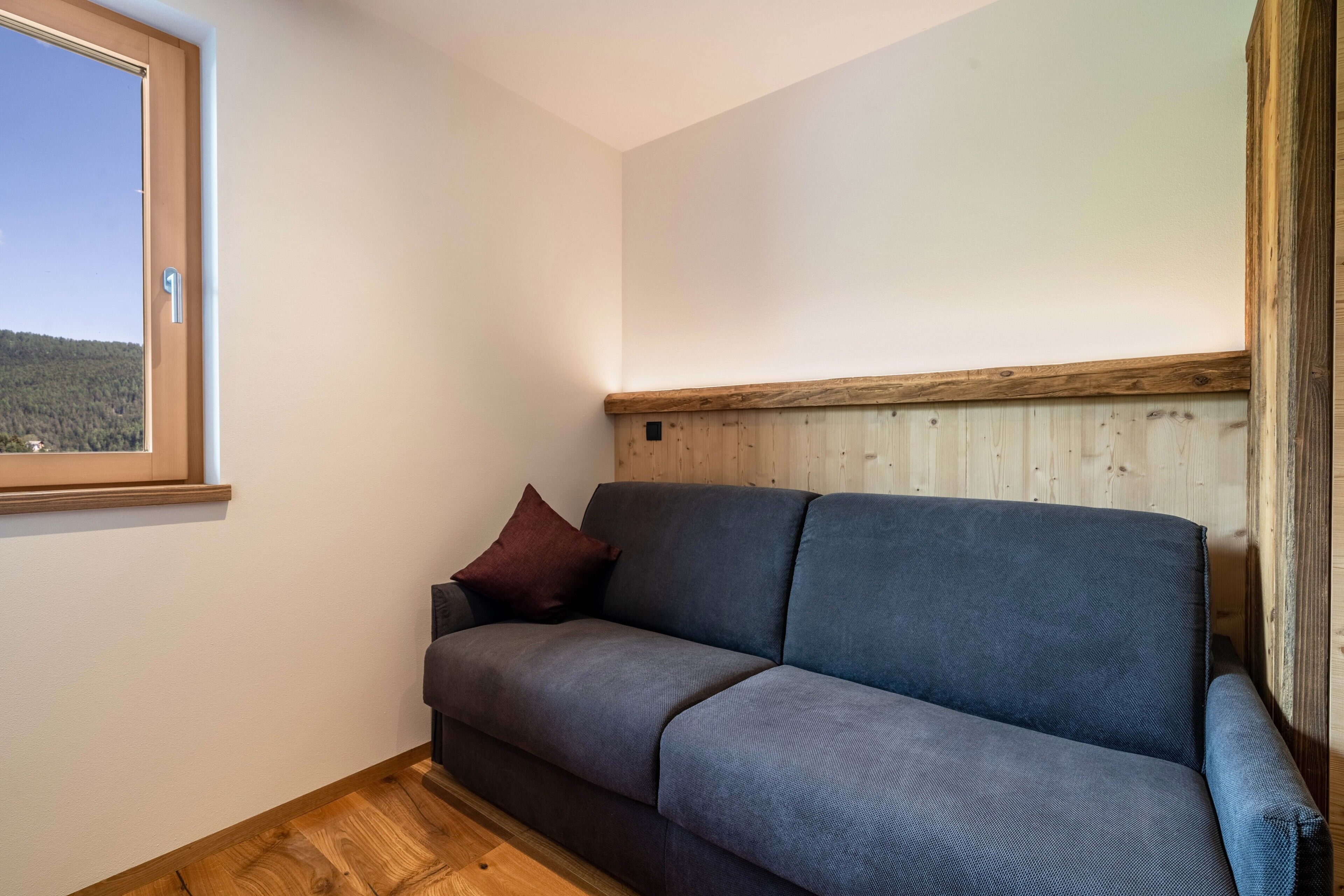 Una habitación con piso de madera, un sofá gris, una ventana y un panel de pared de madera.
