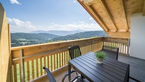 Un balcón de madera con una mesa, sillas, una planta en maceta y vistas a un paisaje montañoso.