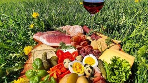 Un montaje para picnic con varios quesos, carnes y verduras sobre una manta, junto con una copa de vino tinto y un paisaje montañoso de fondo.