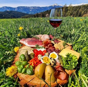 Un pique-nique avec une variété de fromages, de charcuteries et de légumes disposés sur une couverture, accompagné d’un verre de vin rouge et avec une chaîne de montagnes en arrière-plan.