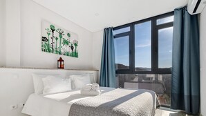 A hotel room with a bed, a window with curtains, a wall-mounted artwork, and a nightstand with a lantern.