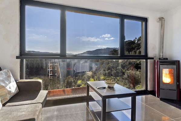 A balcony with a view of a forest and hills, a sofa, a table with a basket, and a wood-burning stove.