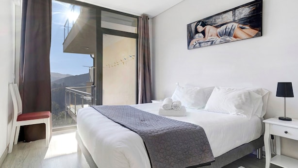 A hotel room with a bed, a painting, a chair, and a view of the outdoors.