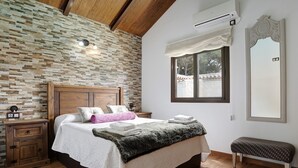 Un dormitorio con pared de piedra, cabecera de madera, cama con ropa blanca, un banco, una mesita de noche y un espejo.