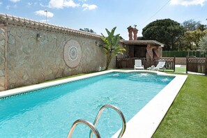 A swimming pool with a stone wall, a small structure with a roof, and a fenced area with lounge chairs.