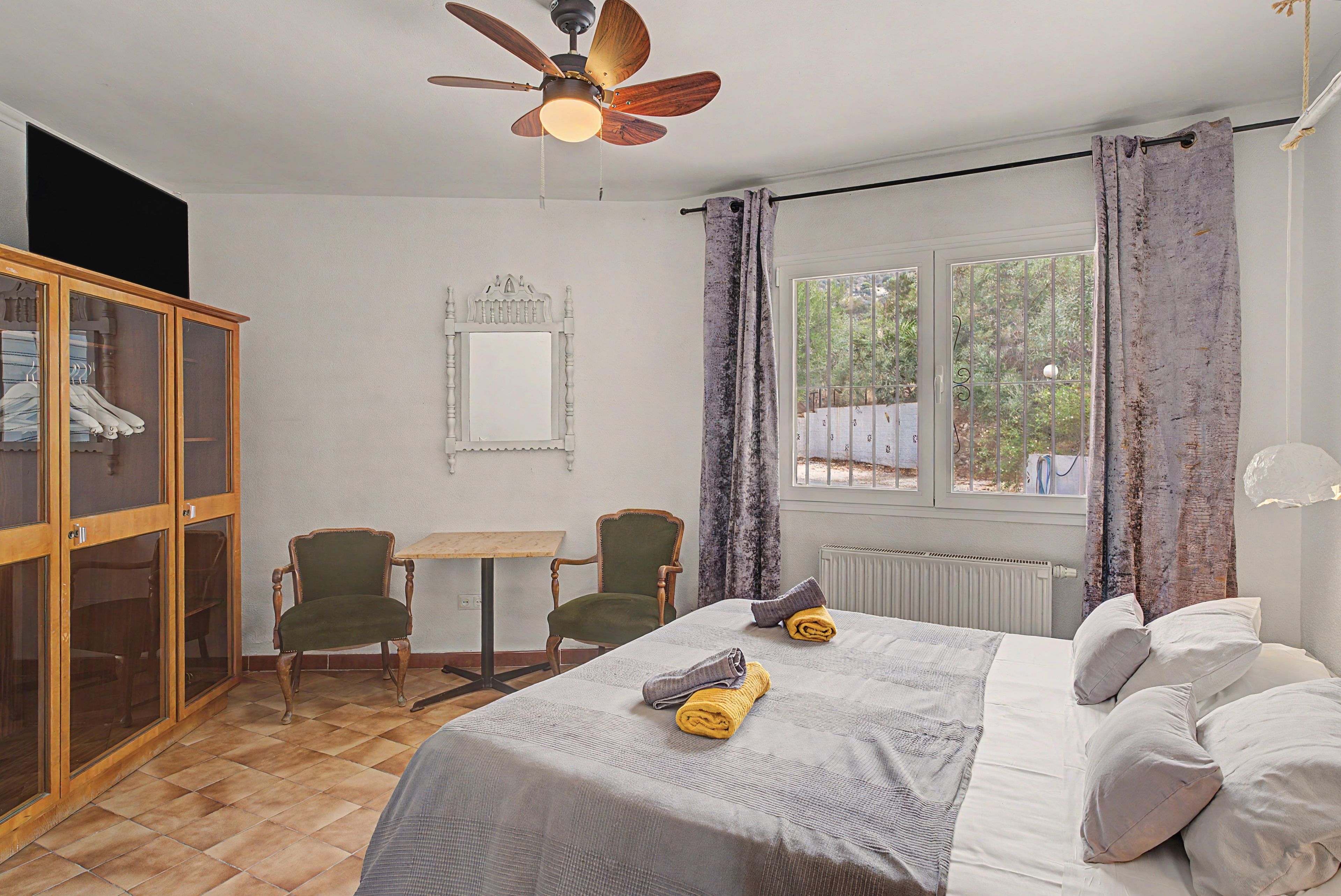A bedroom with a bed, two chairs, a small table, a wardrobe, a ceiling fan, a window with curtains, and a radiator.