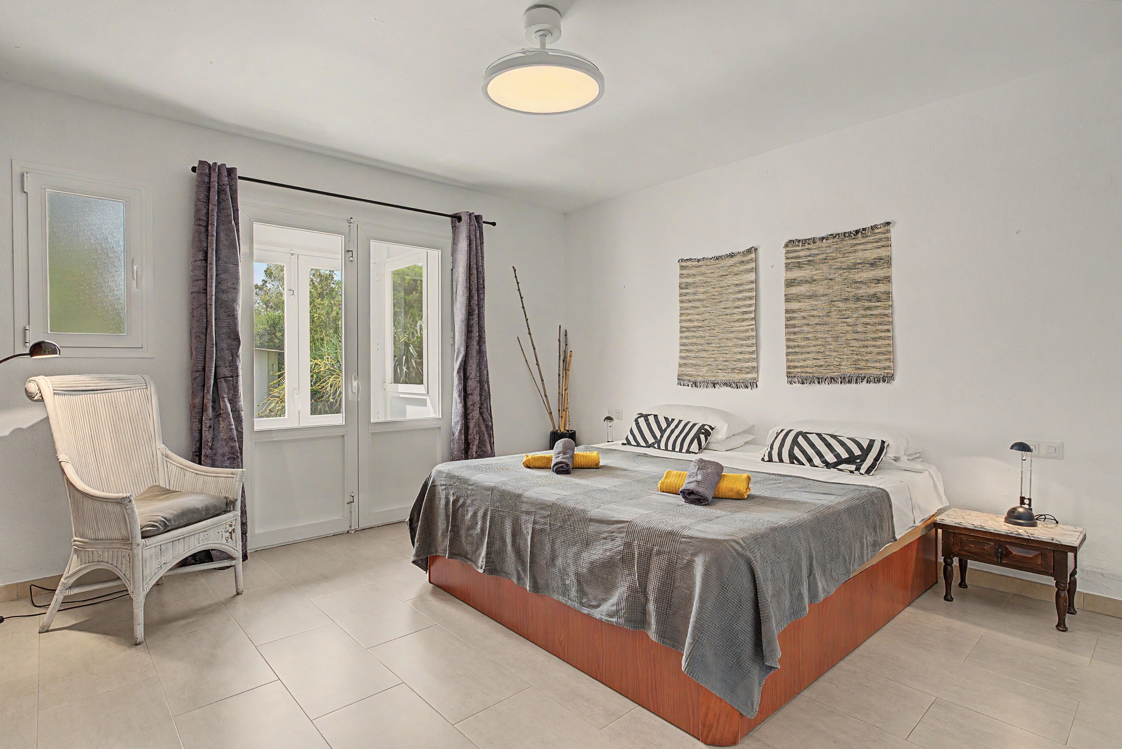 Un dormitorio con cama, una silla, una mesita de noche, una lámpara y un ventanal con cortinas.