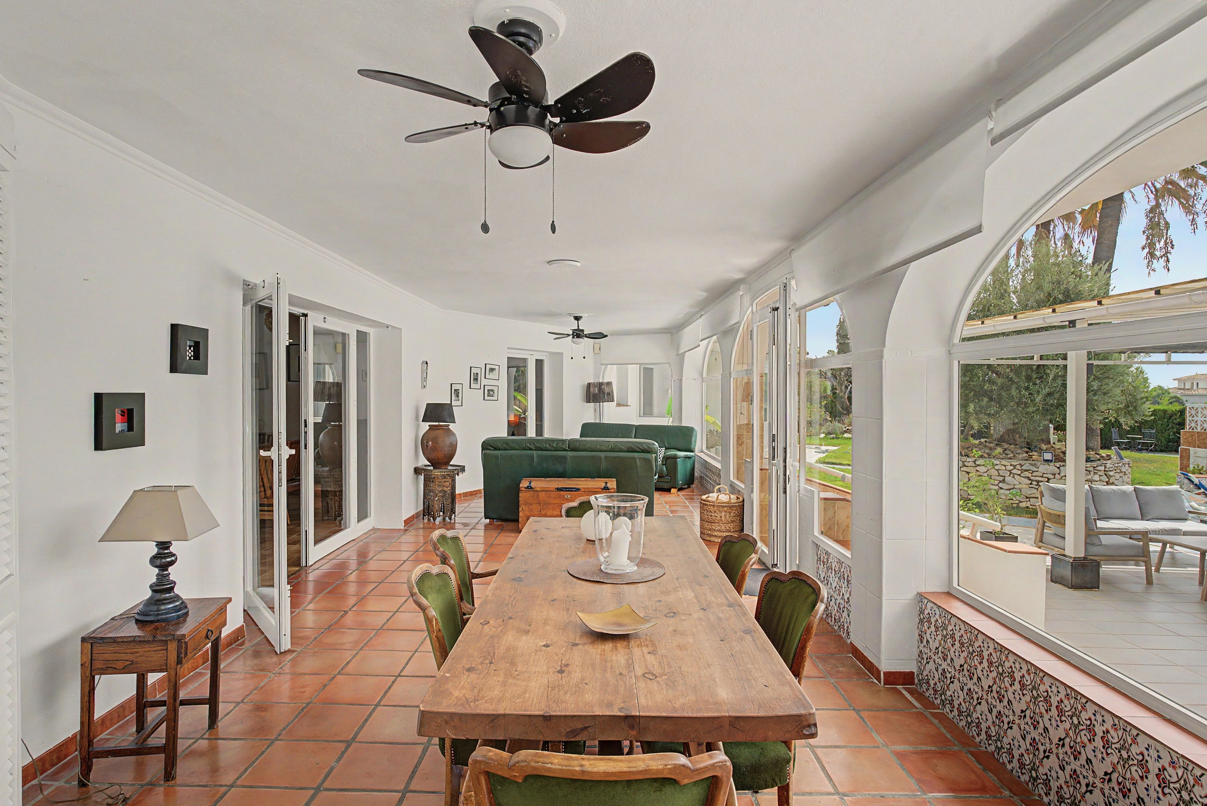 Un vaste espace dédié à la salle à manger, avec une table et des chaises en bois, un ventilateur de plafond et de grandes fenêtres offrant une vue sur l’extérieur.