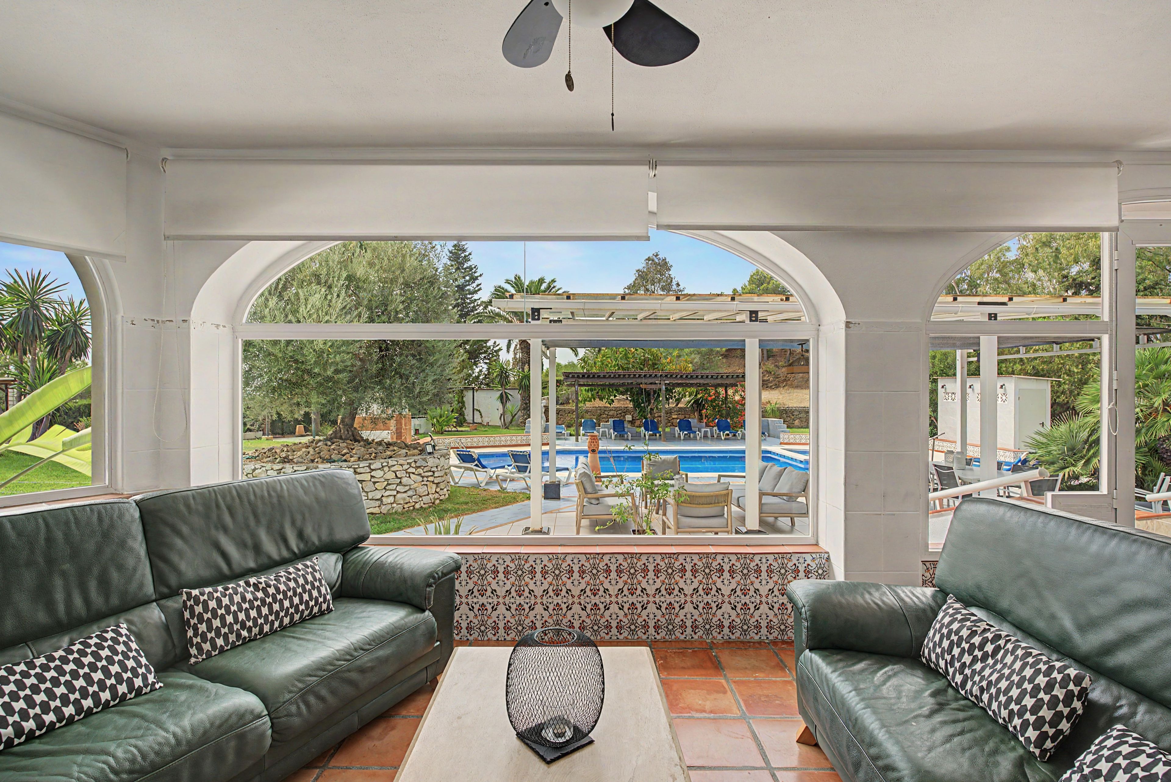Une pièce baignée de lumière, avec deux canapés en cuir vert, une table basse à motifs et une vue sur une piscine et un jardin.