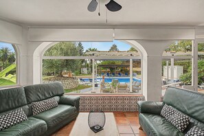 A sunlit room with two green leather sofas, a patterned coffee table, and a view of a swimming pool and garden.