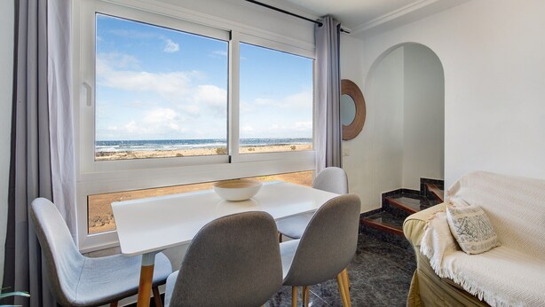 A room with a dining table, two chairs, a sofa, a view of the beach, and a circular mirror on the wall. - Apartment 'El Cotillo Mar Beach' with Sea View, Terrace & Wi-Fi (El Cotillo)