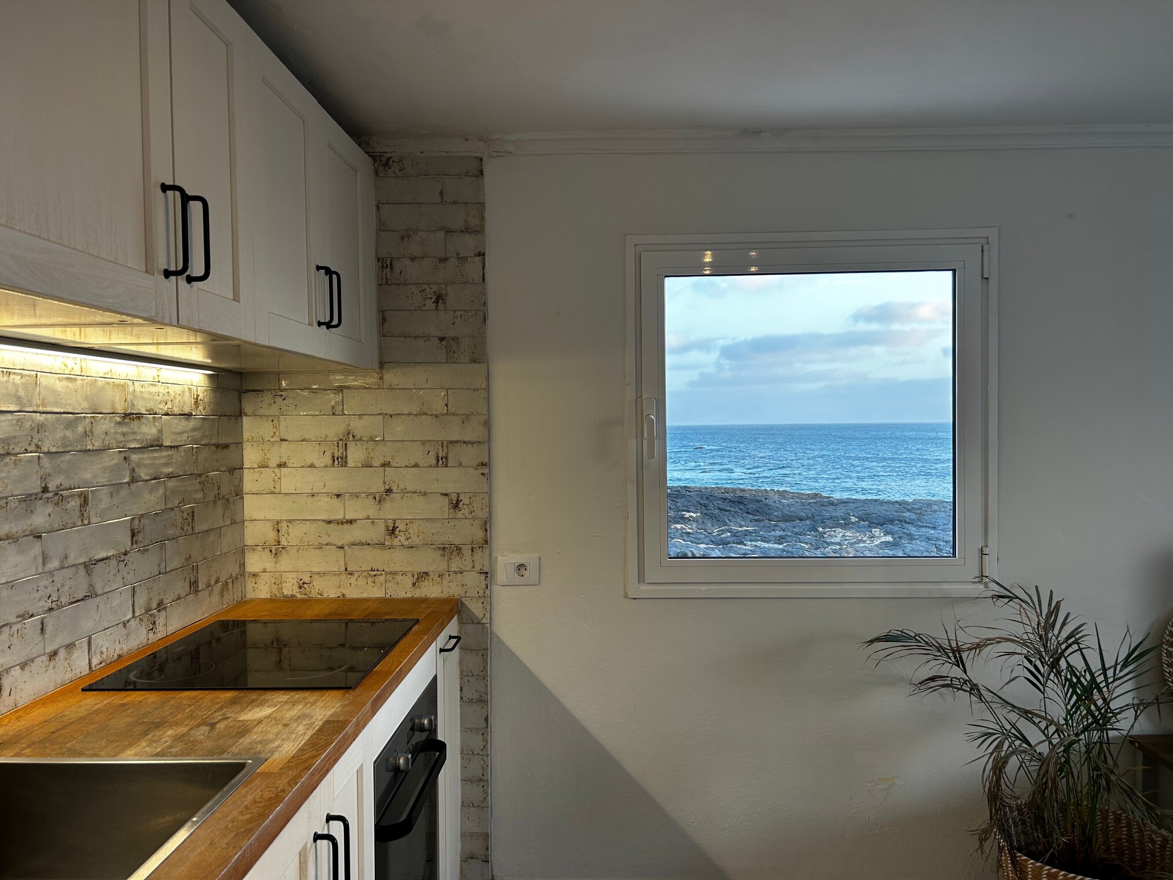 Una cocina con vista al mar a través de una ventana, un muro de ladrillo y encimeras de madera.