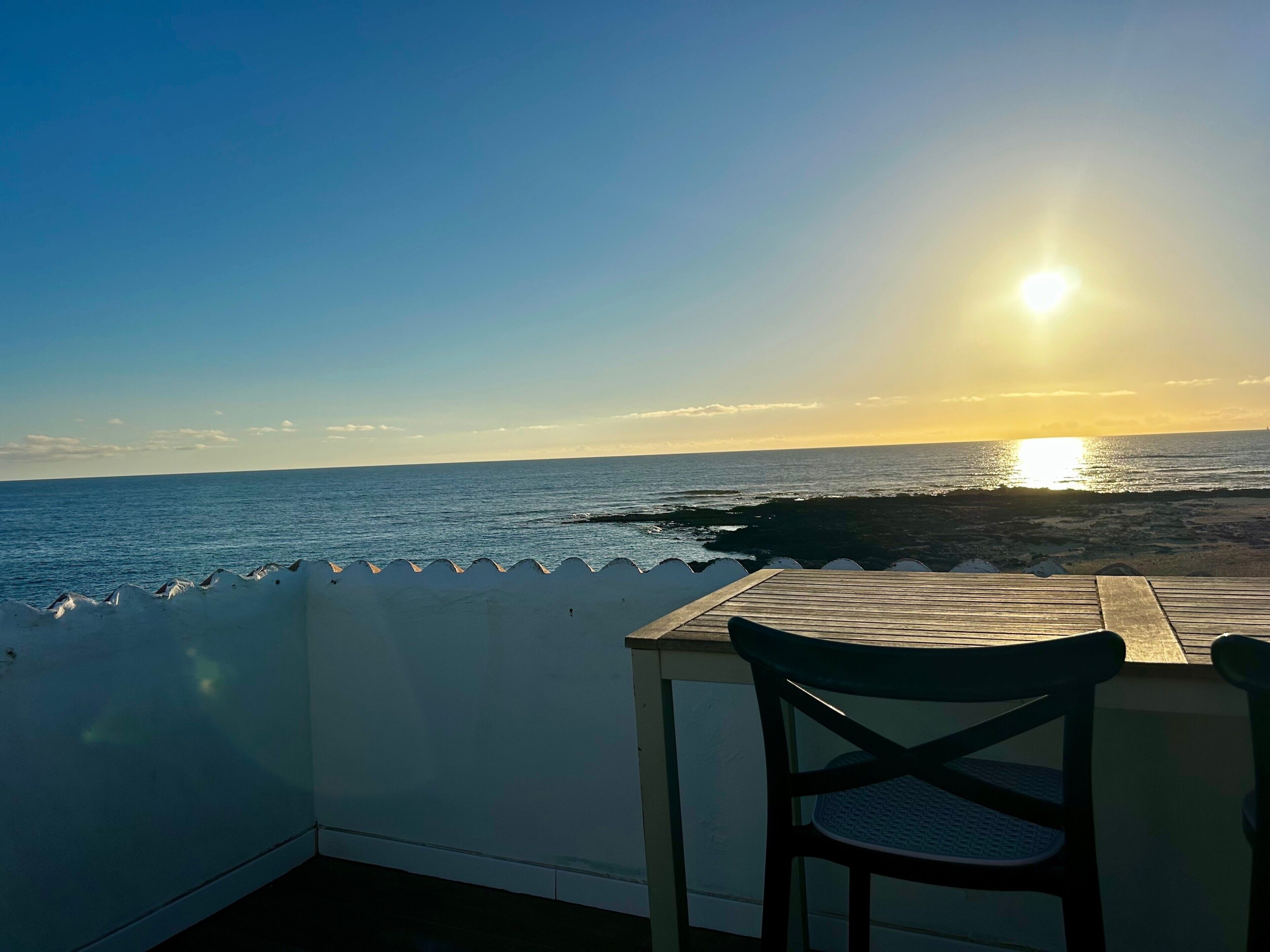 Une table en bois et une chaise sur un balcon donnant sur l’océan, au coucher du soleil.