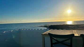 Una mesa de madera y una silla en un balcón con vista al océano al atardecer.