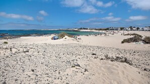 Een strand met rotsachtig terrein, enkele mensen en gebouwen op de achtergrond.