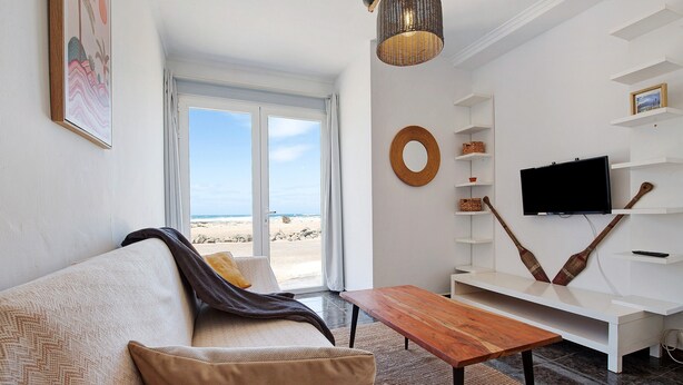 Een moderne woonkamer met een bank, een houten salontafel, een aan een witte muur bevestigde televisie, en een groot raam met uitzicht op het strand.