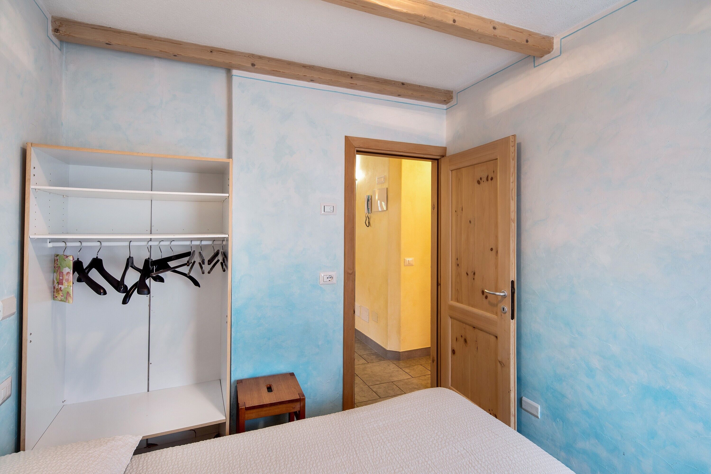A bedroom with a bed, a wooden wardrobe, a small table, and a door leading to another room.